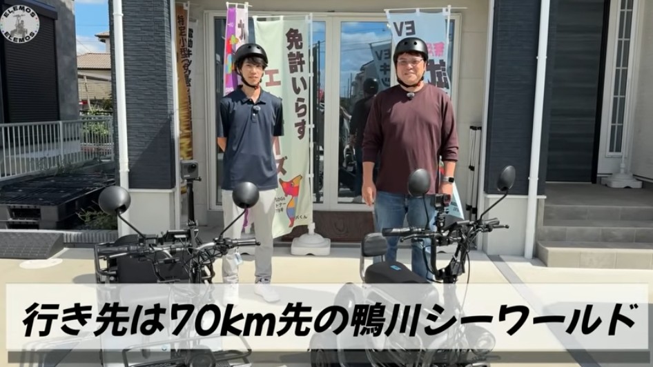 2026-04-21_174613 | ELEMOs Two men in helmets stand beside parked electric bikes in front of a storefront with vertical banners, planning a 70 km ride to Kamogawa Sea World (text banner reads the destination).