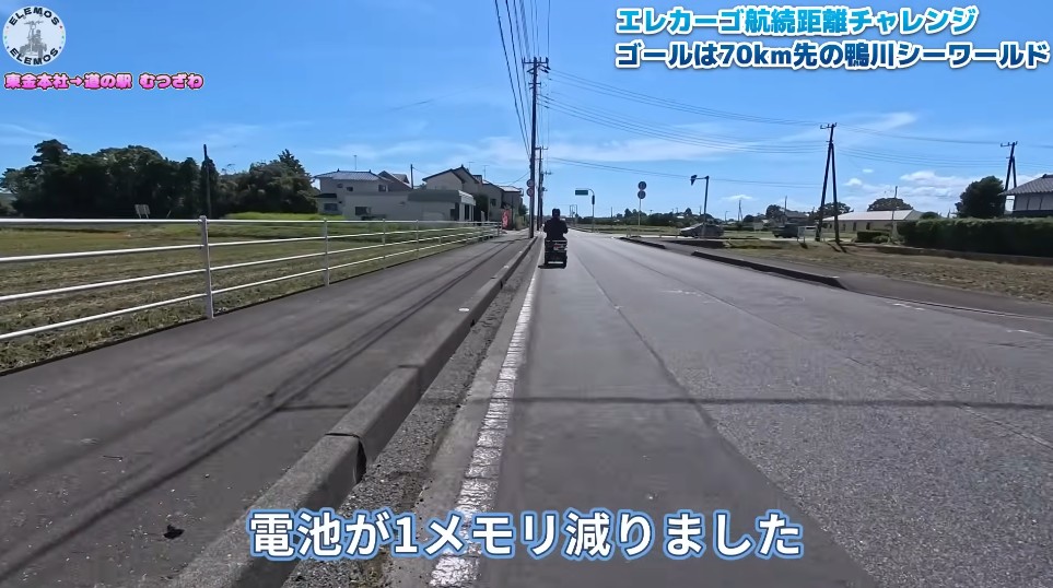 2026-04-21_181828 | ELEMOs Sunny wide suburban road with a scooter ahead; on-screen caption reads '電池が1メモリ減りました' (battery down by one unit).