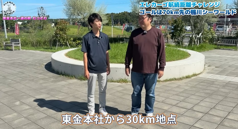 2026-04-21_182346 | ELEMOs Two men stand on a sunny, paved plaza beside a circular planter in a park, talking; a video caption notes they are 30 km from the headquarters.