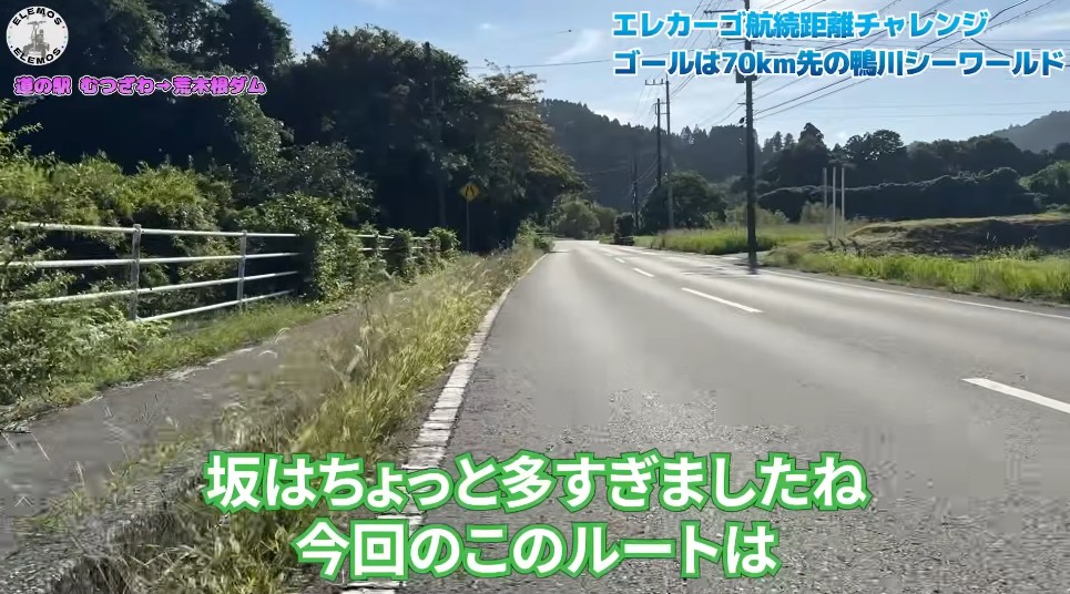 2026-04-21_183043 | ELEMOs Rural road with a grassy verge and trees on the left, utility poles on the right; on-screen Japanese captions reference an electric cargo distance challenge and a 70-km goal.