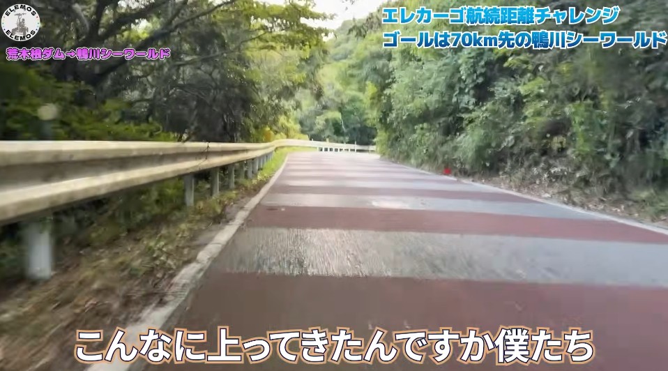 2026-04-21_194900 | ELEMOs Winding mountain road with a guardrail on the left and dense trees on both sides; an overlaid caption at the bottom reads, “こんなに上ってきたんですか僕たち” (Did we climb this much?). Top-right overlay mentions the endurance challenge and 70 km goal.