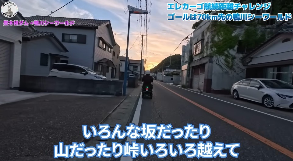 2026-04-21_195256 | ELEMOs Rider on a scooter travels a quiet residential street at sunset; houses, parked cars, and overhead power lines with blue Japanese captions overlaid.