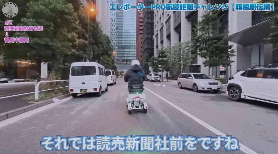 2026-05-04_160832 | ELEMOs Person on a white mobility scooter rides down a city street with tall buildings and parked cars on both sides in daytime urban traffic (video screenshot).