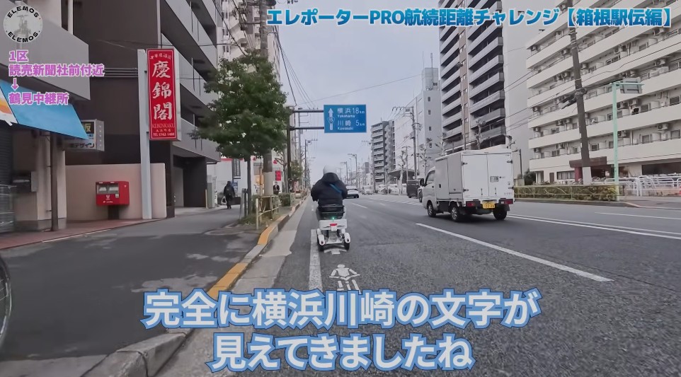 2026-05-04_162711 | ELEMOs 街中を走る二輪車の映像。画面上部に青いタイトル『エレポーターPRO航続距離チャレンジ』、下部に『完全に横浜川崎の文字が見えてきましたね』と字幕が表示されている。周囲にはビルと商店、路上の自転車・車両が見える。