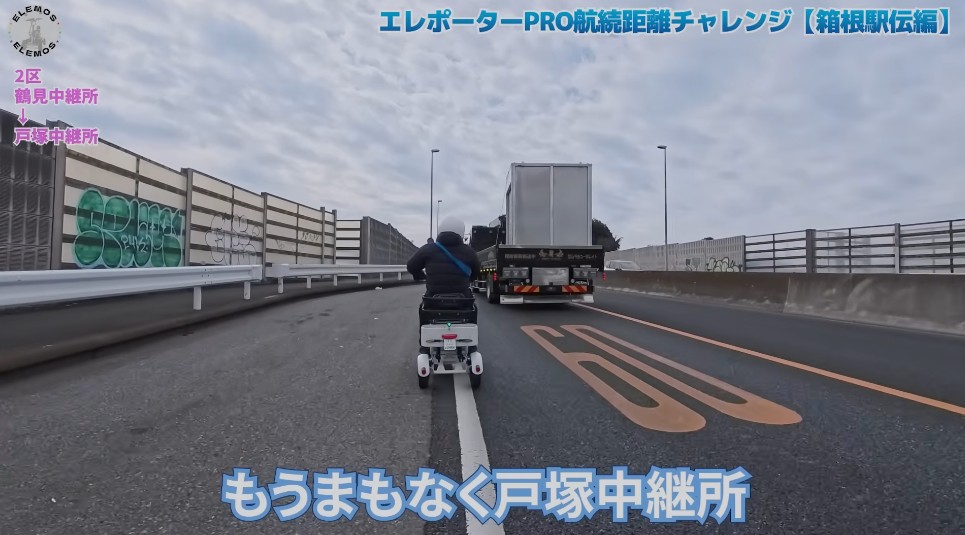 2026-05-04_164758 | ELEMOs Person on a small white electric scooter follows a large truck on a highway, with graffiti-covered barriers and on-screen Japanese video overlays.