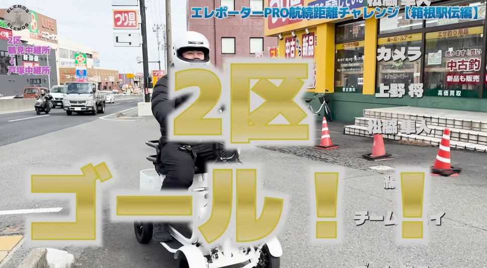 2026-05-04_165112 | ELEMOs Person in a helmet on a white scooter rides toward the camera on a city street, with a large yellow overlay reading '2区 ゴール!!' and storefronts and traffic cones in the background.