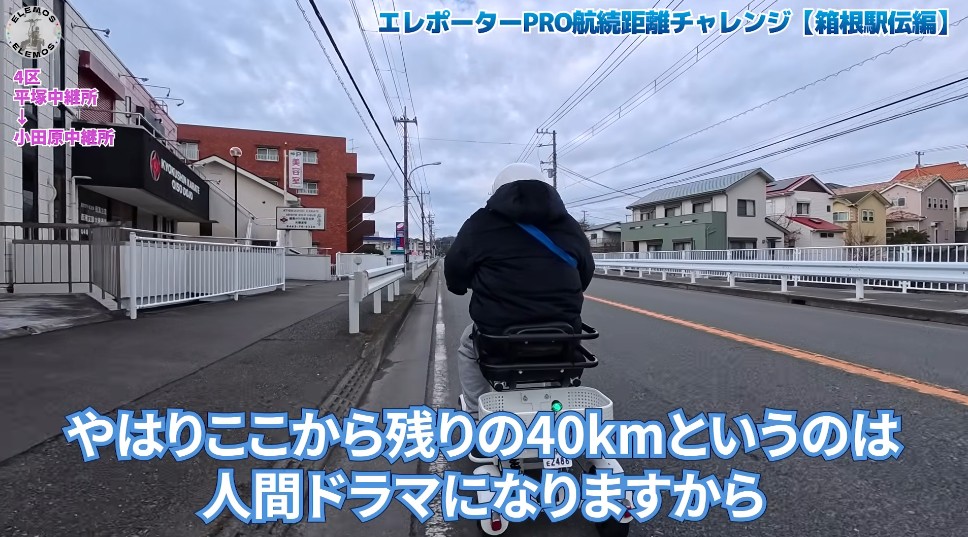 2026-05-04_172527 | ELEMOs Back view of a rider on a small scooter along a suburban street; bottom caption reads 'やはりここから残りの40kmというのは人間ドラマになりますから'.