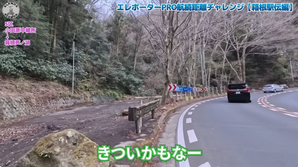 2026-05-04_181717 | ELEMOs Winding mountain road through a forest with a guardrail on the left; two cars drive toward the curve. Signs line the bend.