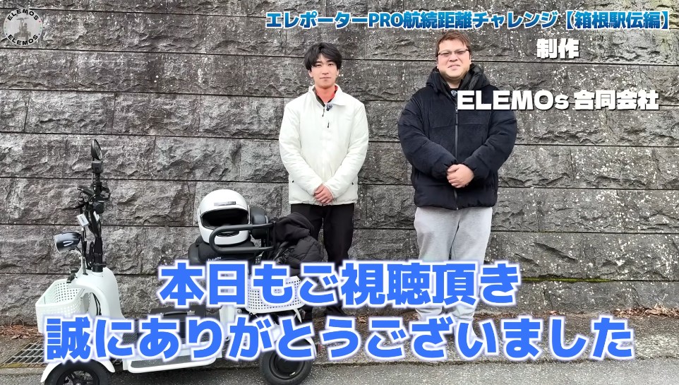2026-05-04_183315 | ELEMOs Two men stand beside a white scooter with a helmet on top, in front of a stone wall; bold blue Japanese text overlays the bottom.