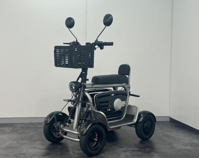 A gray mobility scooter with a front basket, padded seat, and four wheels in a room with a plain wall behind.