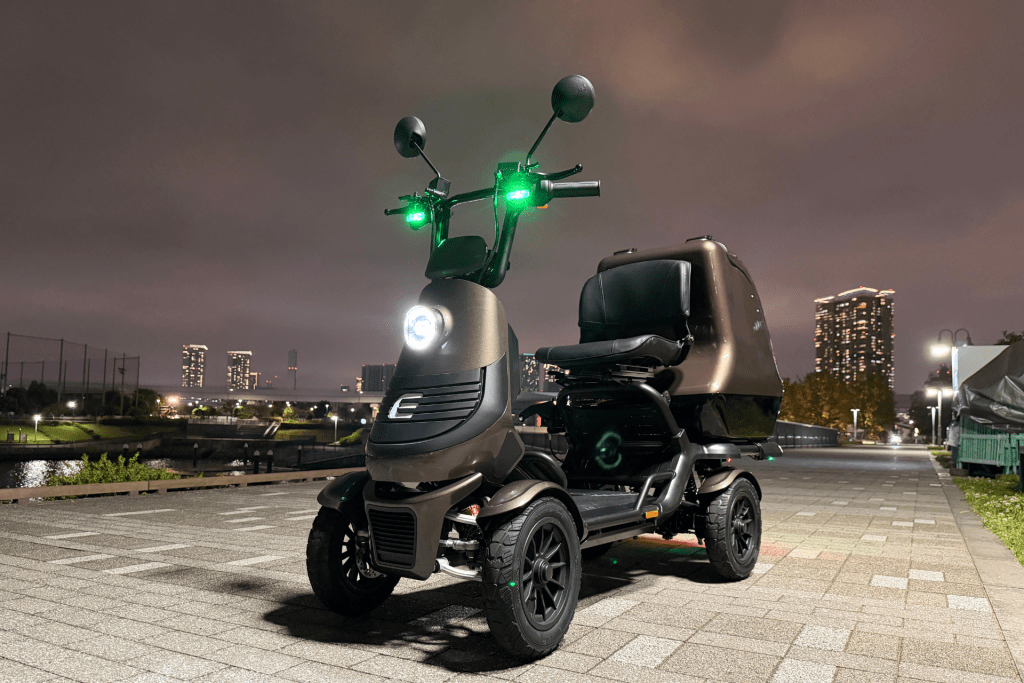 Three-wheeled electric scooter with green front and side lights parked on a paved promenade at night, city skyline in the background