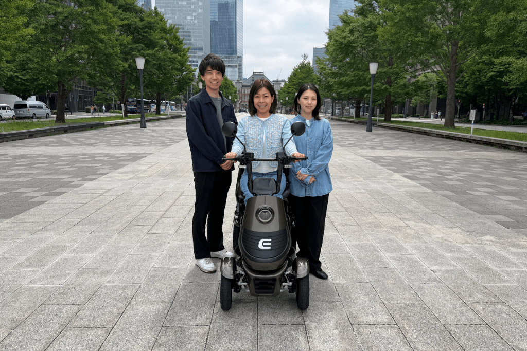 Three friends pose with an electric scooter in a wide city plaza, with tall buildings and green trees behind them.