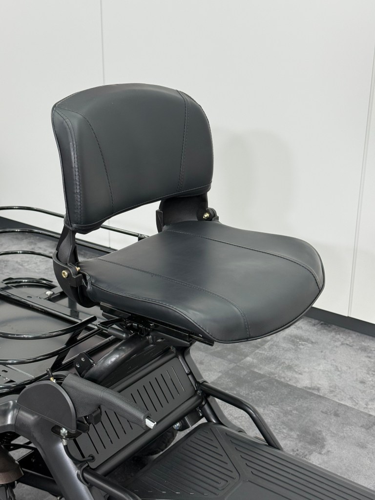 Close-up of a black leather motorcycle/scooter seat with a padded backrest and footboard, indoors on a metal frame.