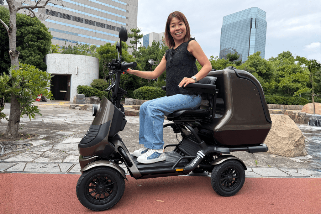Smiling woman sits on a brown mobility scooter outdoors in a plaza with greenery and tall buildings in the background, facing the camera.