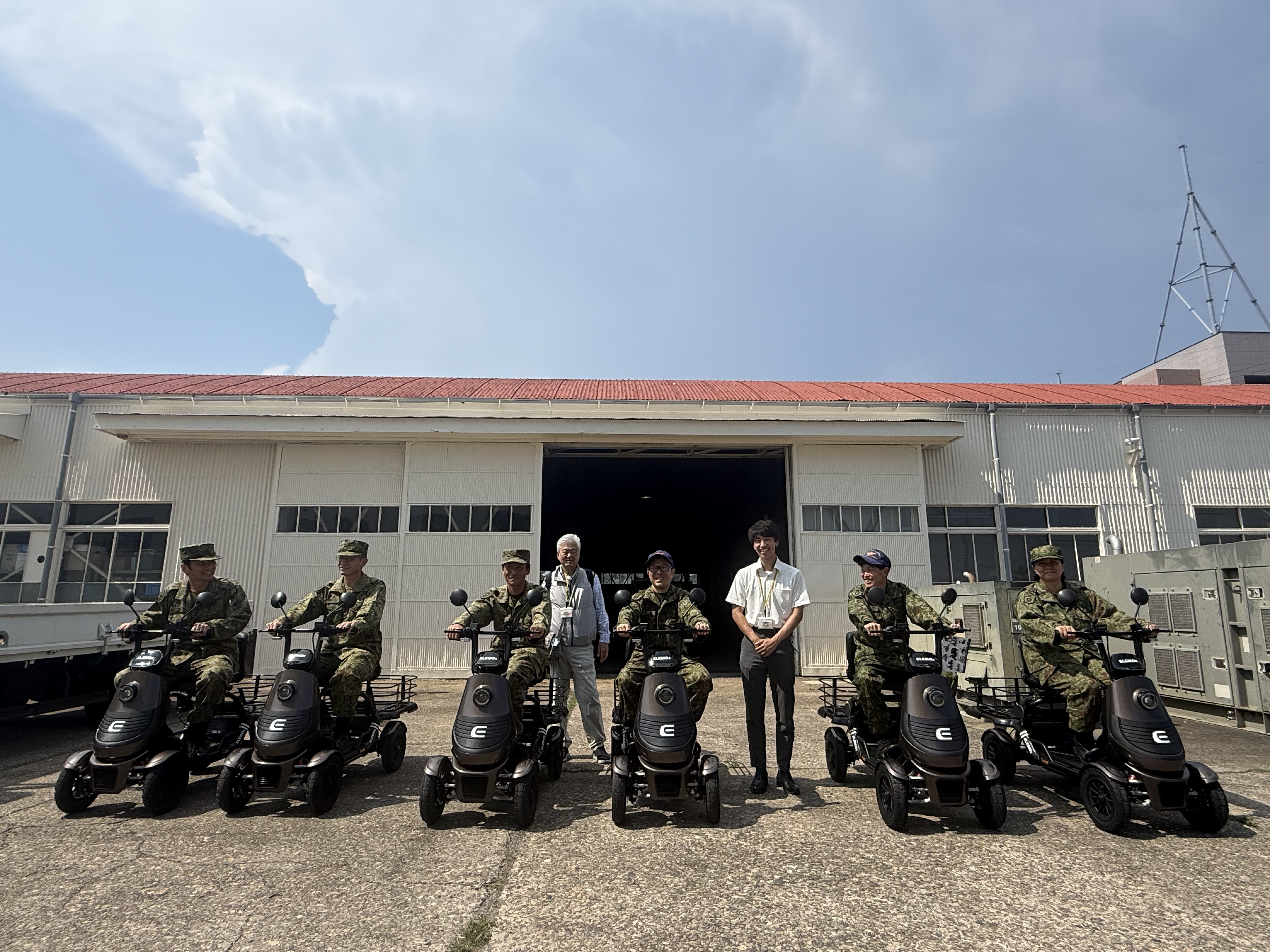 Group of soldiers in uniform posing with a row of mobility scooters outside a warehouse.