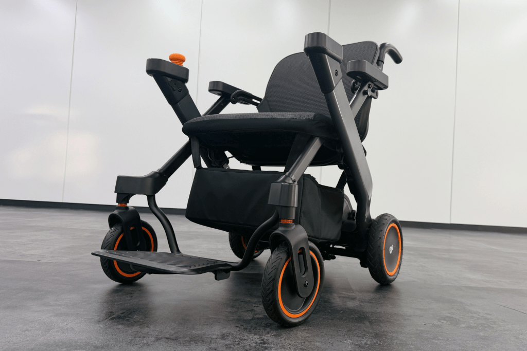 Powered wheelchair with black frame and orange-rimmed wheels, parked indoors on a gray floor.