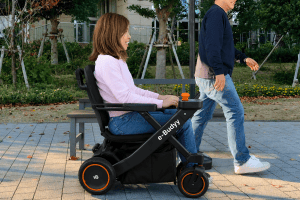 Person in a powered wheelchair being pushed by a companion along a park path.