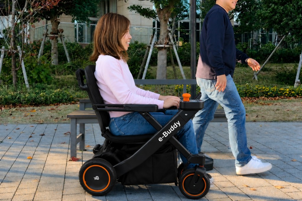 Woman in an electric wheelchair labeled'e-Buddy' sits smiling while a man pushes beside her in a park.
