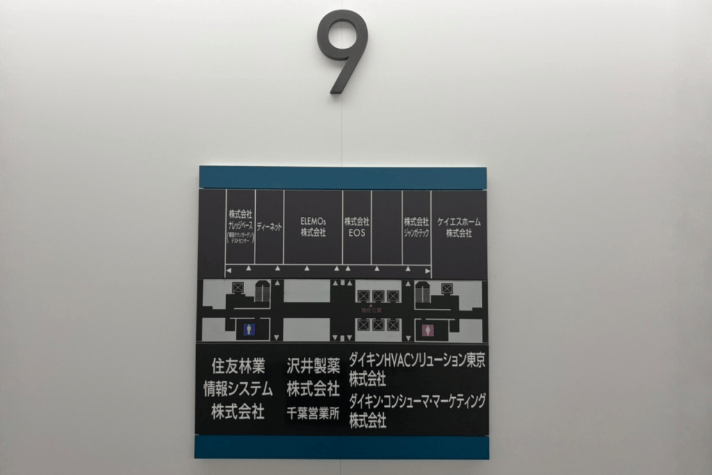 Floor directory sign under a large 9, on a grey wall, listing tenants in Japanese with a simple map layout.
