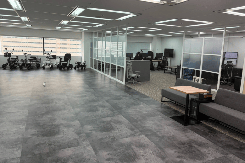 Open office space with several mobility scooters parked by large windows, glass-walled offices in the center, and a gray sofa seating area with a small wooden table on the right side.