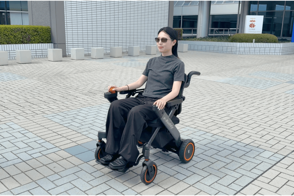 Woman in a black electric wheelchair with orange wheels sits on a tiled outdoor plaza wearing sunglasses and a gray shirt.