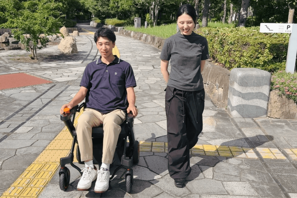Young man in a powered wheelchair on a stone-paved park path, with a smiling woman walking beside him.